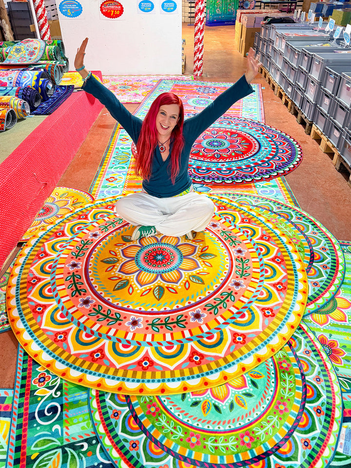 Bine Brändle sitzt lächelnd im Schneidersitz auf einem runden Teppich mit farbenfrohem Mandala-Muster in Gelb, Orange, Türkis, Grün, Pink und Rot und streckt beide Arme nach oben. Um sie herum liegen weitere bunte Teppiche mit Blumen- und Ornamentdesigns in einem Verkaufsraum. Im Hintergrund stehen Teppichrollen, Lagerboxen und dekorative Verkaufsflächen. Die Szene wirkt fröhlich, lebendig und voller kreativer Farben.
