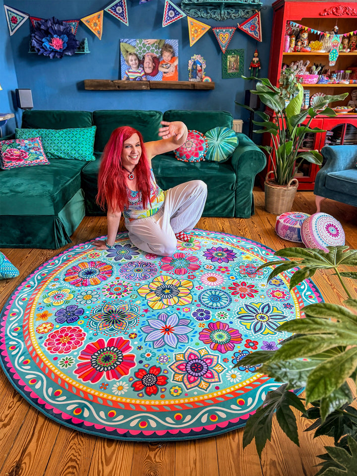 Bine Brändle kniet lächelnd auf einem runden Teppich mit buntem Blumen- und Mandala-Muster in Türkis, Pink, Gelb, Rot, Blau und Lila. Sie hebt eine Hand spielerisch nach oben und befindet sich in einem gemütlichen Wohnzimmer mit grünem Samtsofa, Zimmerpflanzen, bunten Kissen und dekorativen Möbeln vor einer dunkelblauen Wand. Der farbenfrohe Teppich bildet den lebendigen Mittelpunkt der kreativen Boho-Wohnatmosphäre.