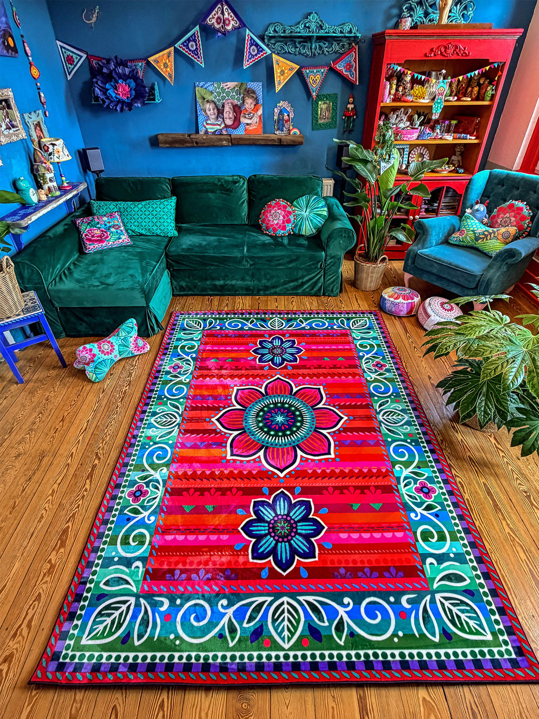 Großer rechteckiger Teppich mit farbenfrohem Blumen- und Ornamentmuster in Rot, Pink, Türkis, Blau, Grün und Lila liegt auf warmem Holzboden in einem gemütlichen Wohnzimmer. Im Hintergrund stehen ein grünes Samtsofa, ein blauer Sessel, Zimmerpflanzen und dekorative Möbel vor einer dunkelblauen Wand mit bunten Girlanden. Der bunte Teppich setzt einen lebendigen Mittelpunkt und schafft eine kreative, wohnliche Boho-Atmosphäre.