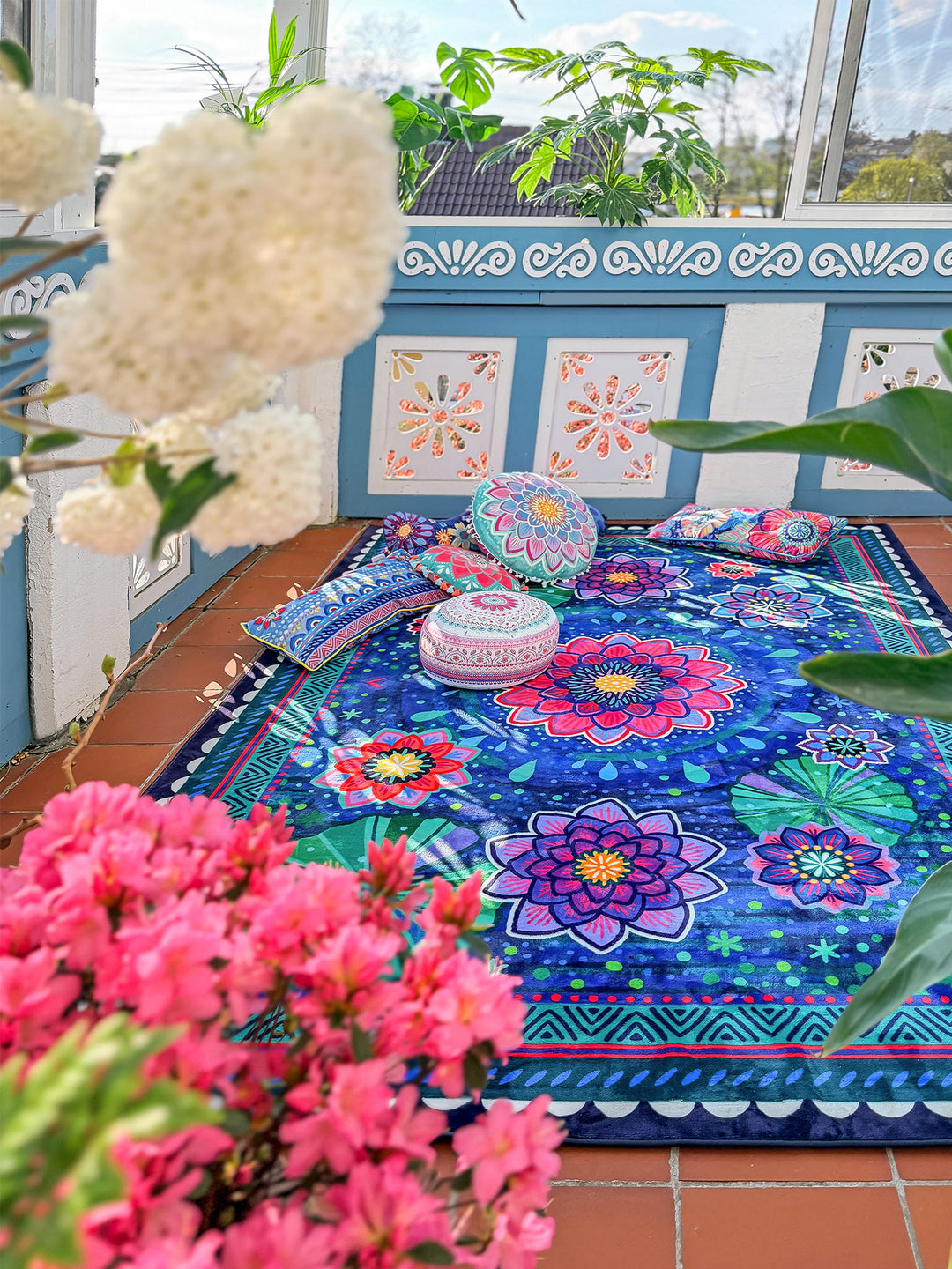 Großer Teppich mit floralem Mandala-Muster in Dunkelblau, Pink, Lila, Türkis und Grün liegt in einem hellen Wintergarten oder auf einer geschützten Terrasse. Auf dem Teppich liegen dekorative Kissen und ein runder Sitzhocker mit bunten Mustern. Im Vordergrund rahmen rosa und weiße Blüten sowie grüne Pflanzen die Szene ein. Sonnenlicht, Pflanzen und farbenfrohe Textilien schaffen eine gemütliche, lebendige Boho-Wohnatmosphäre.