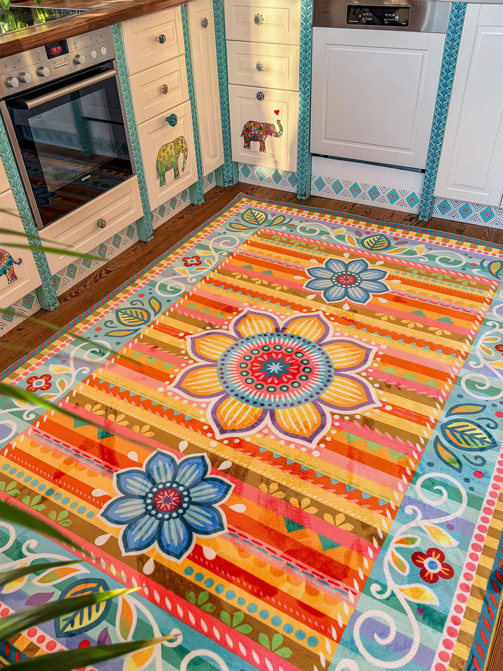 Großer Küchenteppich mit farbenfrohem Blumen- und Ornamentmuster in Orange, Gelb, Türkis, Blau, Rosa und Grün liegt auf Holzboden vor einer hellen Landhausküche. Im Hintergrund stehen weiße Küchenschränke mit dekorativen Elefantenmotiven, Holzarbeitsplatte, Backofen und Geschirrspüler. Der bunte Teppich bringt Wärme, kreative Farben und gemütlichen Boho-Stil in den Küchenbereich.
