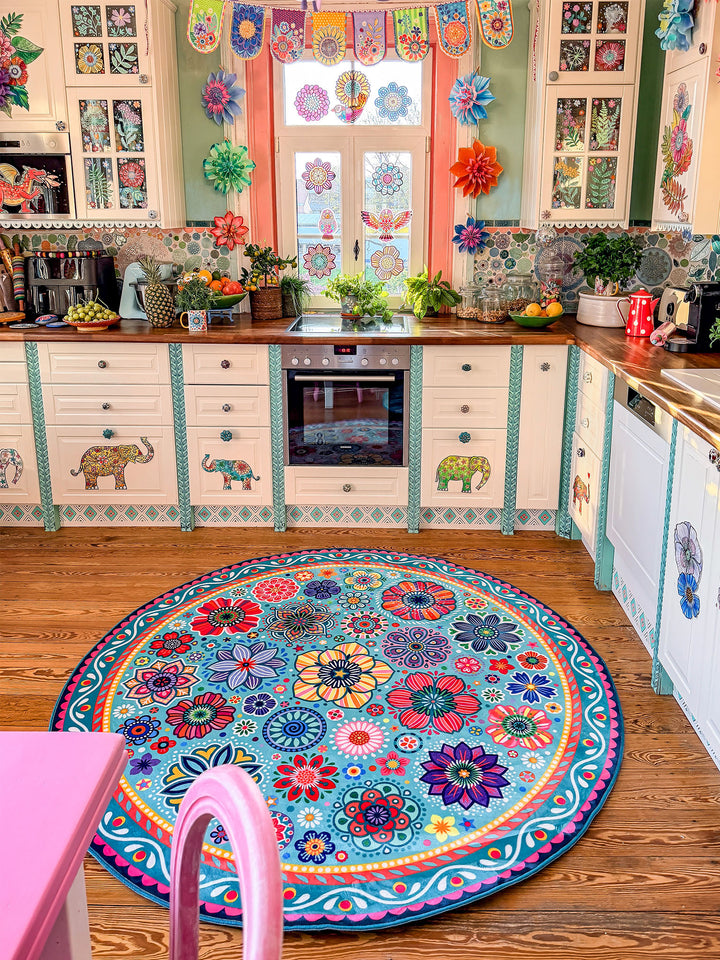 Runder Teppich in Türkis mit farbenfrohen Blumen- und Mandala-Mustern in Pink, Gelb, Rot, Blau, Grün und Lila liegt auf warmem Holzboden in einer kreativ gestalteten Küche. Im Hintergrund stehen helle Küchenschränke mit dekorativen Elefanten- und Blumenmotiven, Holzarbeitsplatten, Pflanzen sowie moderne Küchengeräte. Der bunte Teppich setzt einen lebendigen Mittelpunkt und bringt gemütlichen Boho-Stil in den Küchenbereich.