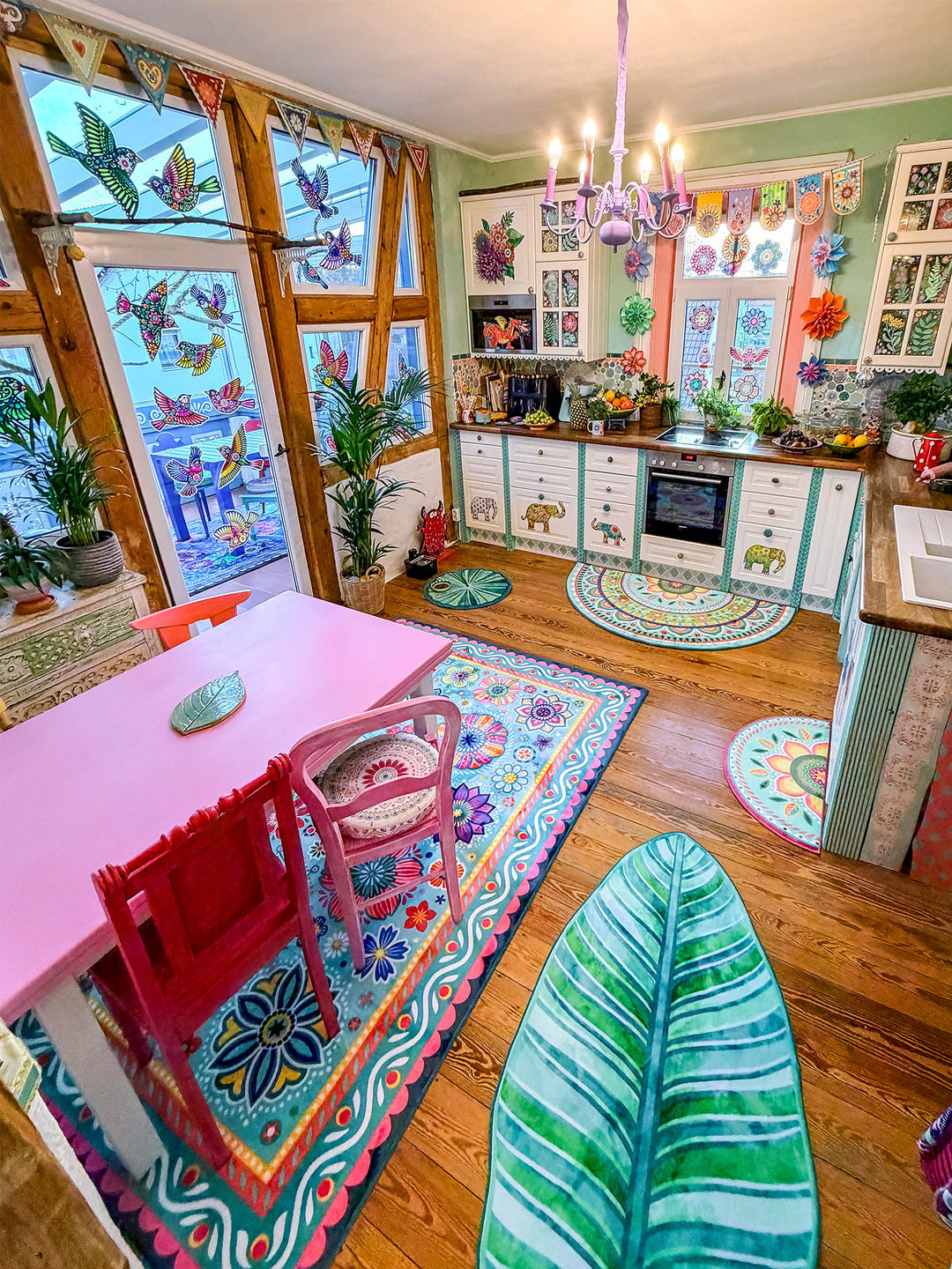 Farbenfrohe Küche im kreativen Boho-Stil mit mehreren dekorativen Teppichen und Matten auf warmem Holzboden. Im Vordergrund liegt eine große blattförmige Matte in Türkis- und Grüntönen, daneben ein rechteckiger Blumenteppich unter einem pinken Esstisch mit bunten Stühlen. Vor der weißen Landhausküche mit Holzarbeitsplatte liegen halbrunde Mandala-Teppiche in Pastellfarben. Große Fenster mit bunten Vogelmotiven, Pflanzen und eine lilafarbene Deckenlampe schaffen eine fröhliche, wohnliche Atmosphäre.