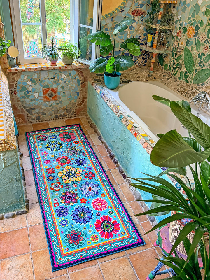 Schmaler rechteckiger Badteppich in Türkis mit farbenfrohen Blumen- und Mandalamotiven in Pink, Blau, Gelb, Rot und Grün liegt auf terrakottafarbenen Fliesen vor einer Badewanne. Das kreative Badezimmer ist mit kunstvollen Mosaikwänden, Naturstein-Details und Pflanzen dekoriert. Große grüne Blätter und ein Fenster mit Tageslicht schaffen eine frische, natürliche Atmosphäre. Der lebendige Teppich setzt einen fröhlichen Farbakzent im individuell gestalteten Boho-Bad.