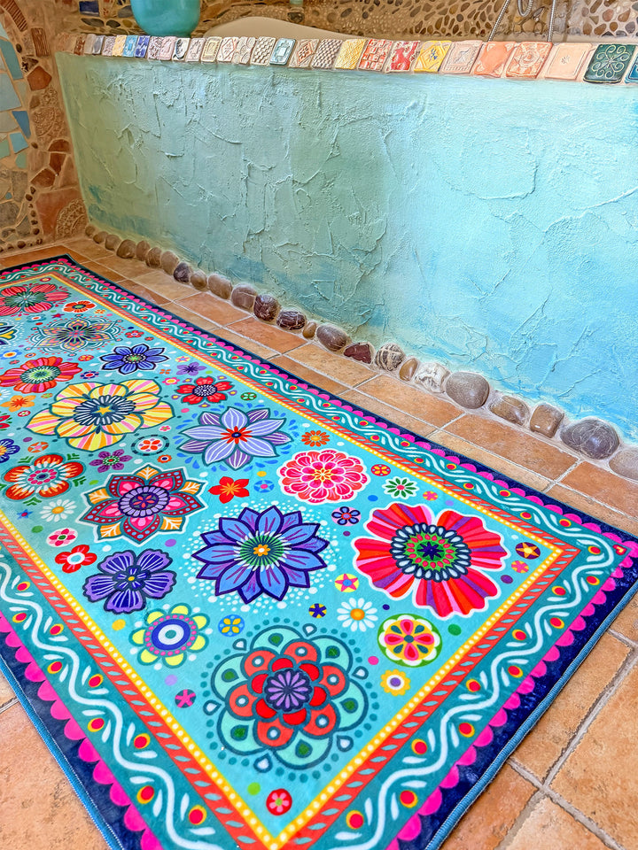 Schmaler rechteckiger Badteppich in Türkis mit farbenfrohen Blumen- und Mandalamotiven in Pink, Blau, Gelb, Rot und Grün liegt auf terrakottafarbenen Fliesen vor einer Badewanne. Das kreative Badezimmer ist mit kunstvollen Mosaikwänden, Naturstein-Details und Pflanzen dekoriert. Große grüne Blätter und ein Fenster mit Tageslicht schaffen eine frische, natürliche Atmosphäre. Der lebendige Teppich setzt einen fröhlichen Farbakzent im individuell gestalteten Boho-Bad.