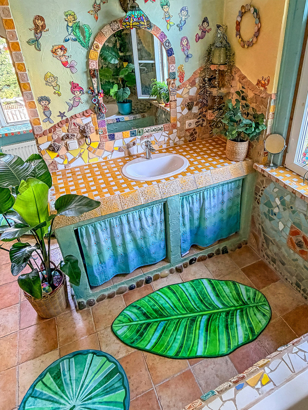 Farbenfrohes Badezimmer im Boho-Stil mit mehreren dekorativen Badteppichen auf warmen Terrakottafliesen. Im Vordergrund liegt ein großer Teppich in Form eines tropischen Blattes mit detailreichen Blattadern in satten Grün- und Limetönen. Dahinter ergänzen ein runder grüner Blätterteppich und ein halbrunder Mandala-Teppich in Türkis, Pink, Gelb und Blau das harmonische Gesamtbild. Links steht eine große Zimmerpflanze im geflochtenen Korb, rechts befindet sich eine Badewanne mit türkis verputzter Front und bu